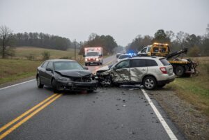 Two damaged cars collide in the middle of a rural road, debris scattered as an emergency tow truck arrives in the background.